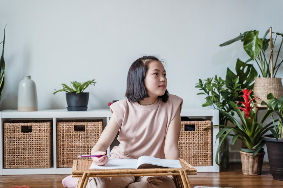 Asian girl focused on homework at a small table, surrounded by indoor plants in a cozy room.