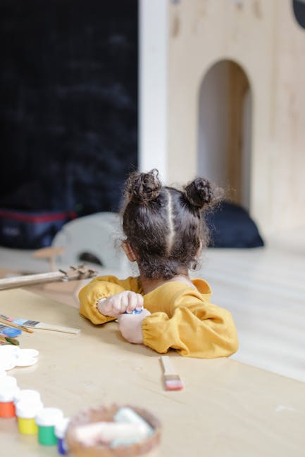 A young girl in a yellow sweater sits indoors, creatively playing at a table with paint.